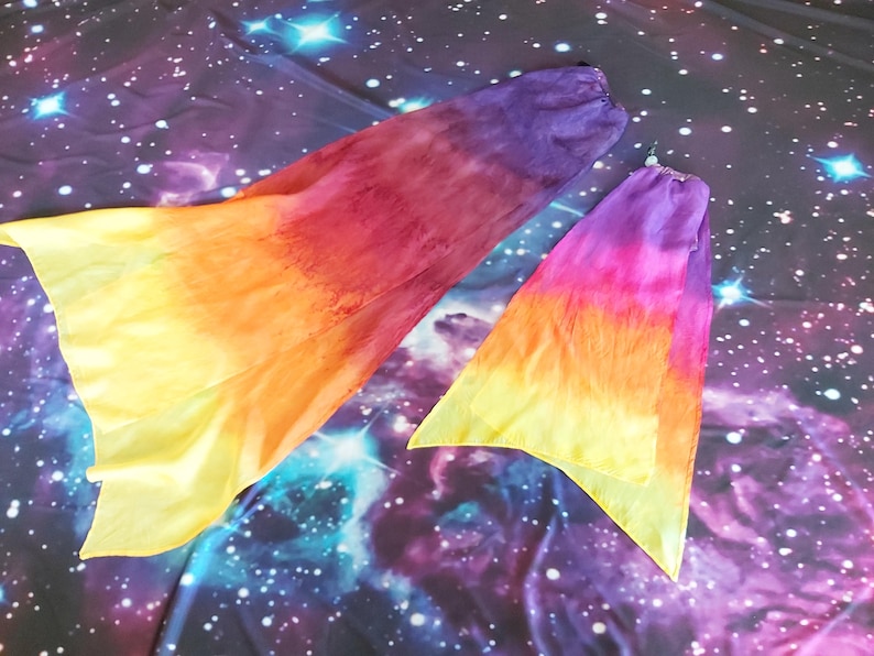 May include: Two flowy fabric dance veils featuring a vibrant gradient pattern that transitions from deep purple at the top to magenta, orange, and bright yellow at the bottom edges. The lightweight material has a soft, semi-sheer texture.