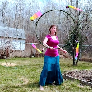 May include: A woman in a pink shirt and a blue skirt is performing with a hoop and colourful ribbons. The hoop is black and has a rainbow ribbon attached to it. The woman is outdoors in a grassy area.
