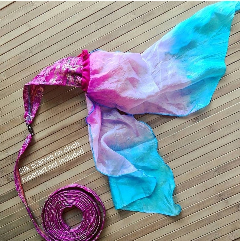 May include: A flowy fabric accessory featuring a vibrant pink and blue gradient silk scarf attached to a patterned magenta fabric cinch. The text "Silk scarves on cinch ropedart not included" is printed on the fabric. The long magenta strap is coiled.