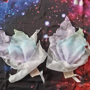 Puede incluir: Dos varitas de tela de color pastel con bordes ondulados. Las varitas están hechas de una tela transparente e iridiscente y están atadas juntas con una cinta blanca.