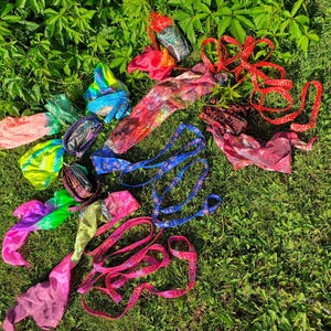 May include: A collection of colourful fabric poi props featuring various patterns and textures. The set includes multiple silk-like streamers in bright neon gradients, blue patterned ribbons, and red fabric strips, all designed for rhythmic movement.