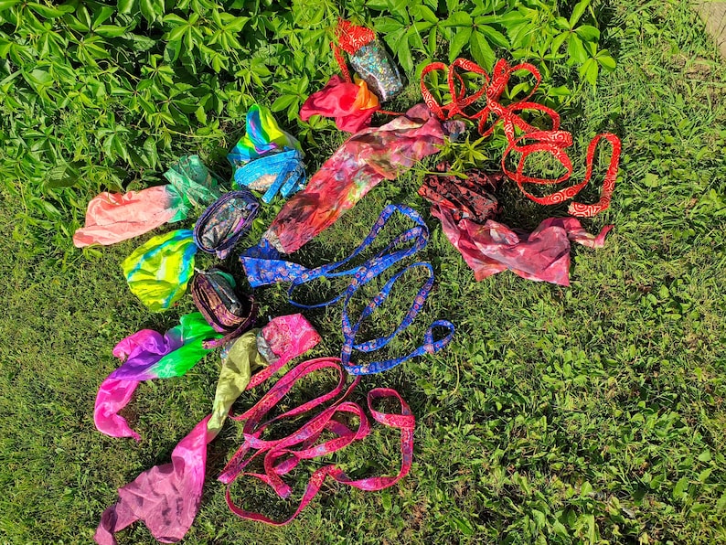 May include: A collection of colourful fabric poi props featuring various patterns and textures. The set includes multiple silk-like streamers in bright neon gradients, blue patterned ribbons, and red fabric strips, all designed for rhythmic movement.