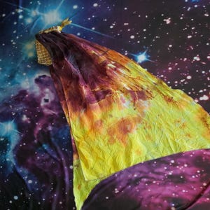 May include: A triangular silk veil featuring a gradient of deep purple and burgundy transitioning into a bright yellow hue. The fabric has a crinkled texture and is attached to a small, gold-toned, mesh-patterned fabric handle at the corner.