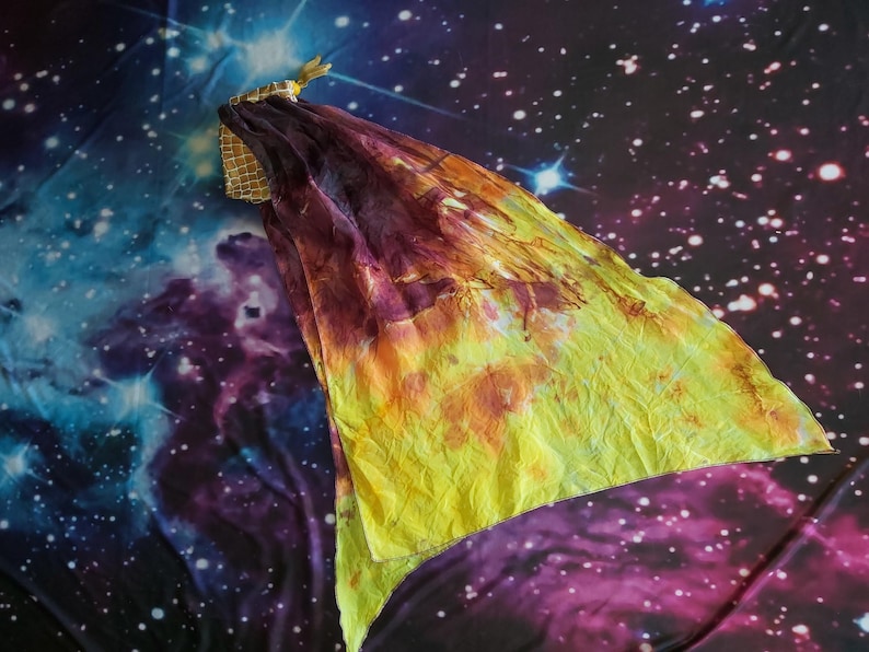 May include: A triangular silk veil featuring a gradient of deep purple and burgundy transitioning into a bright yellow hue. The fabric has a crinkled texture and is attached to a small, gold-toned, mesh-patterned fabric handle at the corner.