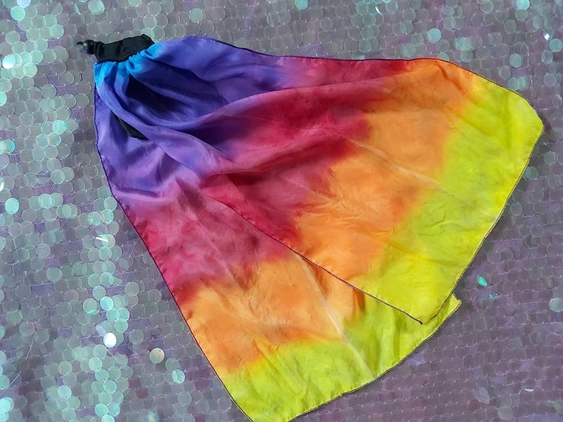 May include: A flowing fabric accessory featuring a vibrant gradient of colours transitioning from deep purple to magenta, orange, and bright yellow. The piece is gathered at one end with a black cord lock mechanism, creating a soft, draped effect.
