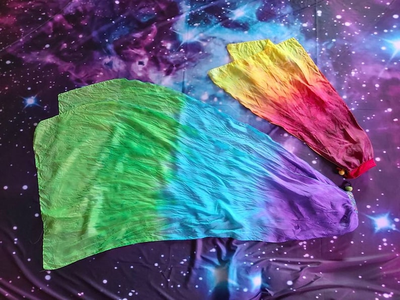 May include: Two dance flags made of lightweight fabric. The larger flag features a gradient of green, blue, and purple. The smaller flag displays a gradient of yellow, orange, and red. Both flags have small, round, coloured beads attached at the base.