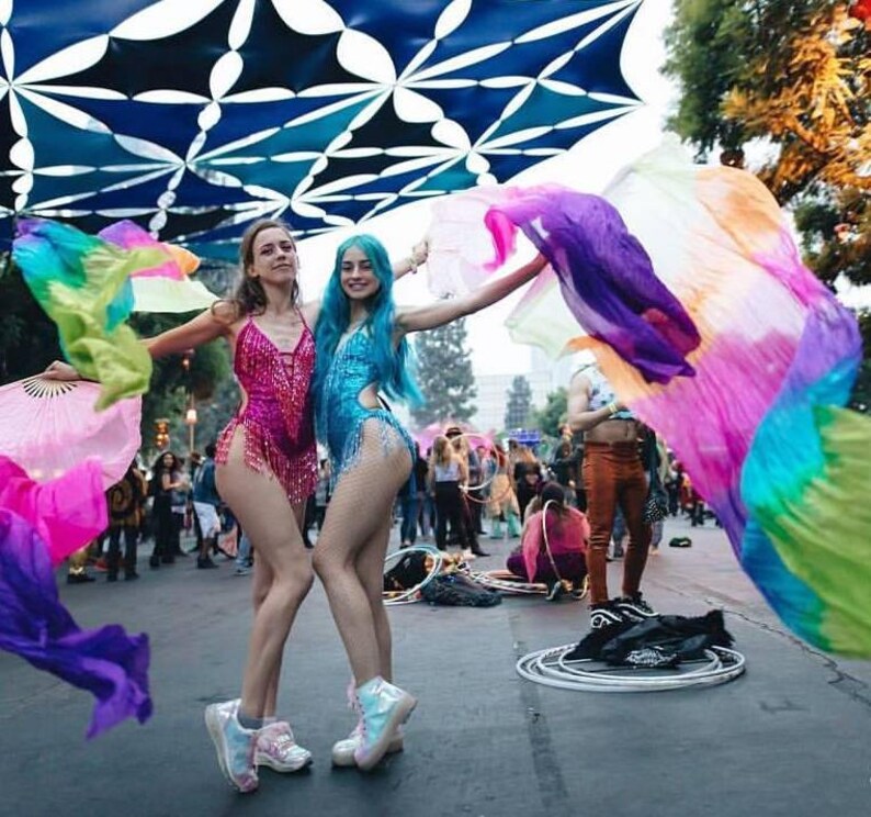 Peut inclure: Deux femmes portant des costumes color&eacute;s et tenant des rubans de tissu, dansent devant une canop&eacute;e &agrave; motifs bleus et blancs. Les femmes portent des v&ecirc;tements color&eacute;s et ont les cheveux longs. L'arri&egrave;re-plan est une foule de personnes.