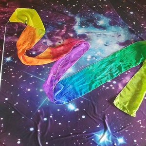 May include: A rainbow colored silk ribbon streamer with a white stick attached to one end. The streamer is lying on a purple and black fabric with a galaxy print.