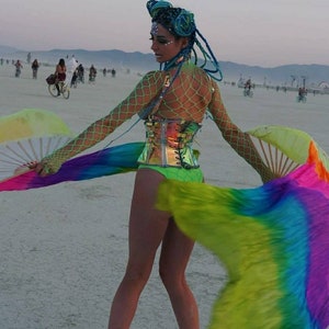 Peut inclure: Une femme portant un corset vert et argent&eacute; avec un haut en r&eacute;sille et des extensions de cheveux bleus. Elle tient des &eacute;ventails arc-en-ciel et se tient dans un paysage d&eacute;sertique.