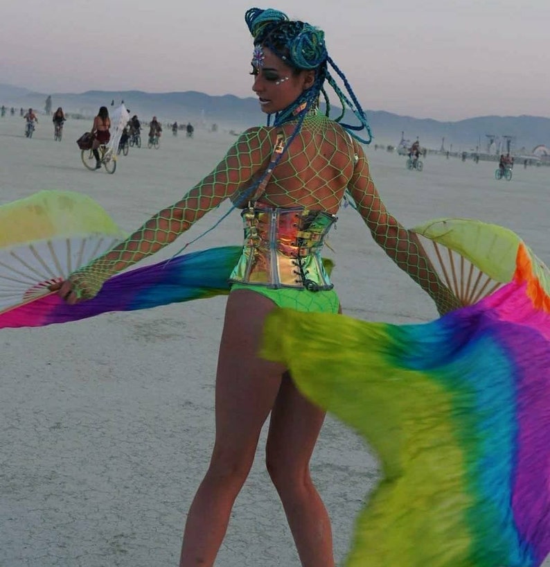 Peut inclure: Une femme portant un corset vert et argent&eacute; avec un haut en r&eacute;sille et des extensions de cheveux bleus. Elle tient des &eacute;ventails arc-en-ciel et se tient dans un paysage d&eacute;sertique.