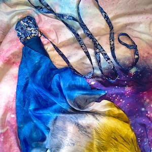 May include: A long, flowing silk scarf featuring a vibrant gradient of blue, white, and yellow. The scarf is attached to a dark blue strap with a repeating silver dragon pattern, which includes a loop at the end for handling or attachment.
