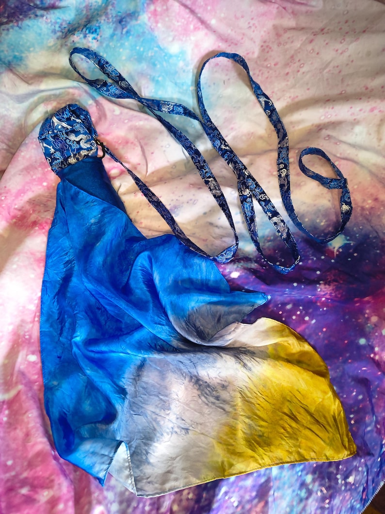 May include: A long, flowing silk scarf featuring a vibrant gradient of blue, white, and yellow. The scarf is attached to a dark blue strap with a repeating silver dragon pattern, which includes a loop at the end for handling or attachment.