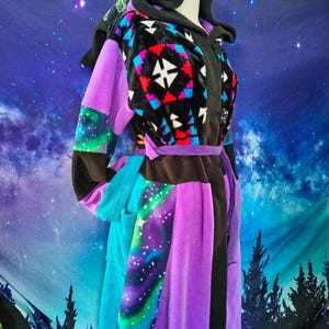 Puede incluir: Una bata con capucha de patchwork con una paleta de colores vibrantes. La bata presenta un diseño negro, morado y verde azulado con un patrón geométrico en el pecho. Las mangas y el cuerpo tienen un estampado de galaxia y paneles morados lisos. Un cinturón a juego ciñe la cintura.