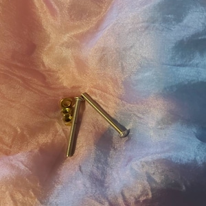 Puede incluir: Dos tornillos plateados y cuatro tuercas doradas sobre un fondo de tela de color rosa y azul degradado.