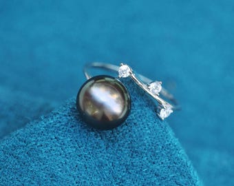 May include: A silver ring with a large, dark gray pearl and three small, clear gemstones. The ring is set against a teal blue background, creating a striking contrast. The pearl has a lustrous sheen.
