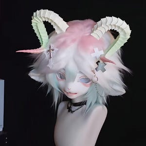 May include: A fantasy-themed headpiece with white, pink, and green accents. The headpiece features large, curved horns, fluffy white hair, and a stylized face. A cross pendant and chain add detail. The piece is displayed on a mannequin head.