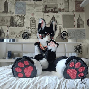 Może przedstawiać: Czarny i biały fursuit z czerwonymi akcentami, z dużymi podkładkami na łapy z różowymi i czerwonymi detalami. Fursuit ma białą klatkę piersiową i szczegółową głowę z goglami. Tło to ściana pokryta znaczkami o tematyce podróżniczej.