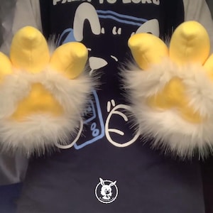 May include: A pair of yellow and white furry hand paws with four fingers each. The paws are worn by a person wearing a blue shirt with a cartoon design.