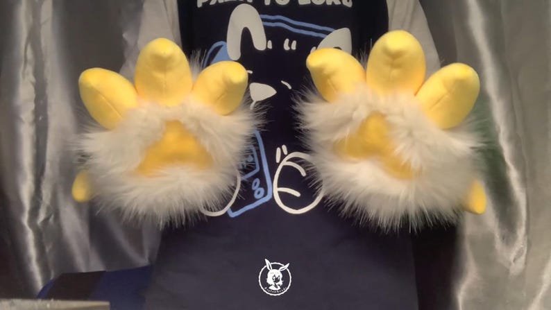 May include: A pair of yellow and white furry hand paws with four fingers each. The paws are worn by a person wearing a blue shirt with a cartoon design.