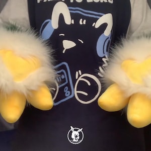 May include: A pair of white and yellow furry hand puppets with three yellow claws on each hand. The puppets are being worn by a person wearing a blue shirt with a cartoon character on it.
