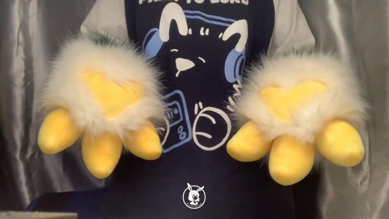 May include: A pair of white and yellow furry hand puppets with three yellow claws on each hand. The puppets are being worn by a person wearing a blue shirt with a cartoon character on it.