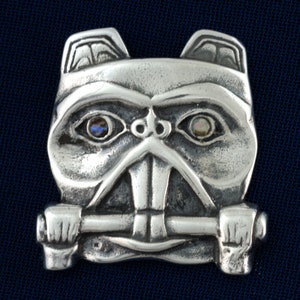 Può includere: Un ciondolo d'argento con un viso stilizzato dettagliato con due occhi opalescenti. Il viso tiene una barra d'argento in bocca.