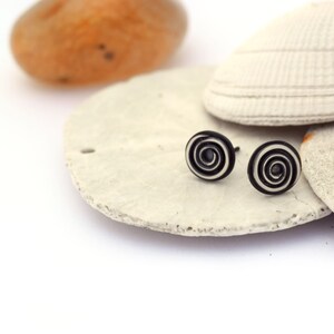 A pair of stud earrings sit on a stack of sea shells with an orange agate in the background. The circular studs have a tight wire spiral. Black oxidation in the depressed areas brings out the spiral. The raised areas have a brushed finish.