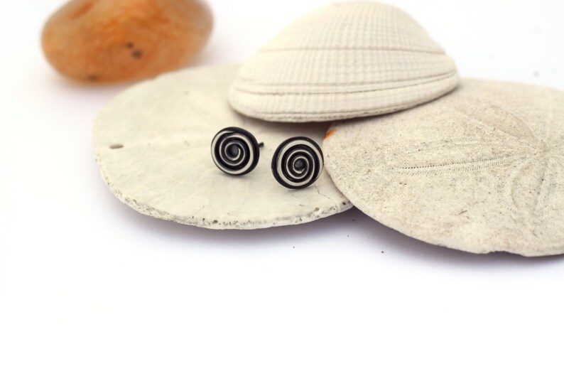 A pair of stud earrings sit on a stack of sea shells with an orange agate in the background. The circular studs have a tight wire spiral. Black oxidation in the depressed areas brings out the spiral. The raised areas have a brushed finish.