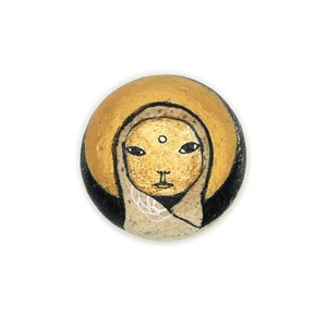 Puede incluir: Una piedra redonda pintada con un fondo dorado y una ilustración en blanco y negro de una cara humana. La persona tiene un halo blanco y un tercer ojo.