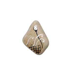 Può includere: Una pietra beige con un disegno dipinto in bianco e nero di un viso stilizzato con uno stelo e foglie. Le foglie sono dipinte di bianco e lo stelo è nero.
