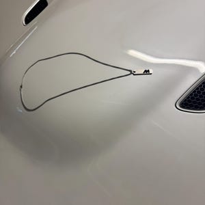 Könnte beinhalten: Silberfarbene Halskette mit einem rechteckigen Anhänger mit dem Buchstaben "M" und einem Streifendesign. Die Kette ist filigran und der Anhänger klein. Die Halskette wird auf einer weißen Oberfläche präsentiert.