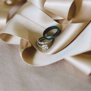 May include: Two wedding bands on a beige satin ribbon. The band on top is a dark blue, while the band underneath is a silver band with small diamonds.