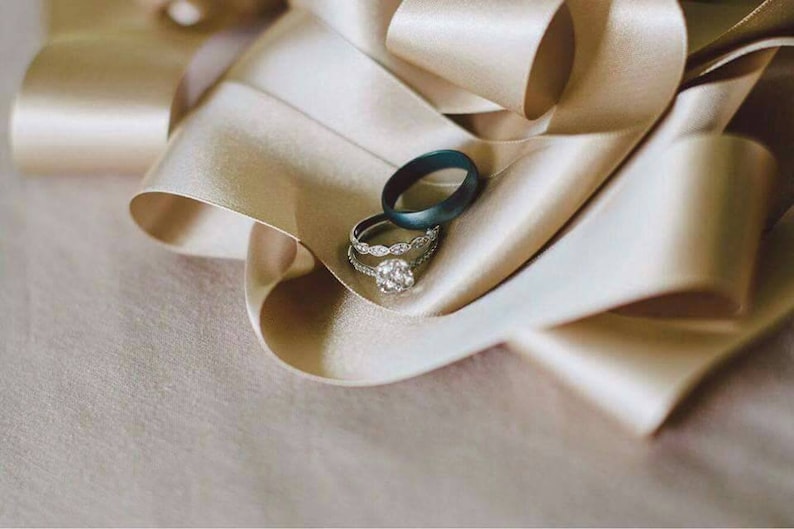 May include: Two wedding bands on a beige satin ribbon. The band on top is a dark blue, while the band underneath is a silver band with small diamonds.