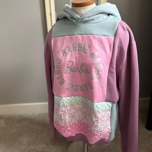 May include: A patchwork hoodie in shades of purple, pink, and blue, displayed on a mannequin. The hoodie features the text "Barbie Camp" in a circular design on the front. The bottom section has a patchwork design.