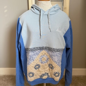 Puede incluir: Una sudadera con capucha Adidas azul claro con un panel estampado floral. La sudadera tiene una capucha con cordón y mangas largas. El logotipo de Adidas está bordado en el pecho. El panel floral presenta diseños florales beige y azules.