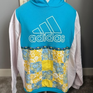 May include: A turquoise hoodie with white sleeves and a patchwork design. The front features the Adidas logo in white. The patchwork includes yellow and blue floral patterns. The hoodie is displayed on a mannequin.