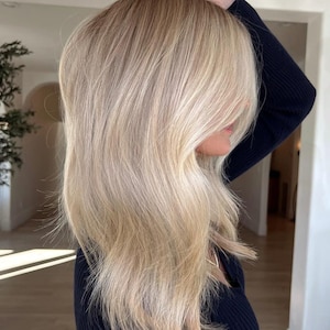 May include: A person with shoulder-length, layered blonde hair featuring bright platinum highlights and darker blonde lowlights. The hair is styled straight with soft volume and a side-swept fringe, worn with a black long-sleeved ribbed shirt.
