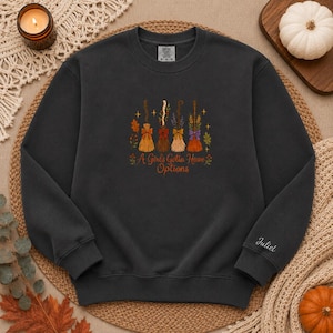 Puede incluir: Una sudadera gris oscuro de cuello redondo con un gráfico de cuatro escobas estilizadas con lazos y follaje de colores. Debajo de las escobas, el texto dice A Girl's Gotta Have Options. La manga izquierda tiene bordado el nombre Juliet.