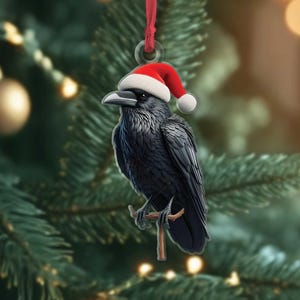 Puede incluir: Adorno navideño de un cuervo negro con un gorro de Papá Noel, posado en una pequeña rama. El adorno está suspendido por una cinta roja sobre un fondo de un árbol de Navidad decorado con bolas y luces doradas.