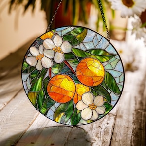 May include: Round stained glass panel featuring oranges, white blossoms, and green leaves. The artwork is framed in black and hangs from a chain. The design is reminiscent of a citrus tree, with vibrant colors and intricate details.