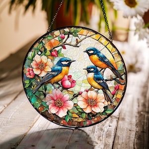Puede incluir: Arte de ventana de vidriera redonda con tres pájaros azules y naranjas posados en ramas florecidas. La obra de arte incluye flores rosas y naranjas, hojas verdes y un marco de metal negro con una cadena para colgar. El fondo es una mezcla de colores.