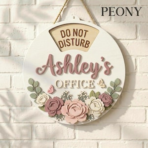 Op de afbeelding: Een ronde, witte houten plaat met de tekst "DO NOT DISTURB" en "Ashley's OFFICE". Versierd met roze en witte bloemen, groene bladeren en vlinders. Opgehangen aan een jute touw.