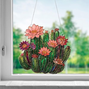 May include: A stained glass cactus window hanging with green cacti and pink and red flowers. The cactus is suspended by a silver chain. The background is a blurred view of green trees and a blue sky.