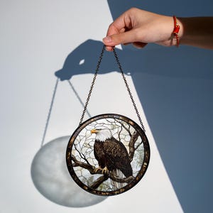 May include: Circular stained glass window depicting a bald eagle on a tree branch. The eagle has a white head and brown body. The window is framed with a dark metal chain for hanging. The background is a light blue and white.