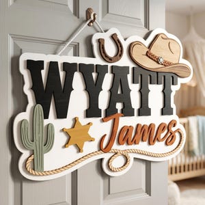 May include: A wooden sign with the name "Wyatt James" in black and brown lettering, featuring a cowboy hat, horseshoe, cactus, and sheriff's badge. The sign is white with a rope border and hangs from a white rope.