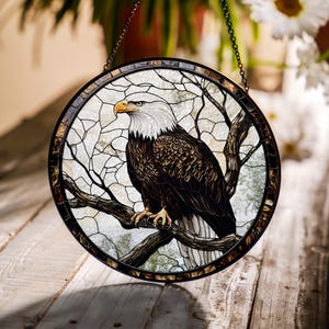 May include: Circular stained glass window depicting a bald eagle on a branch. The eagle has a white head and brown body, with a yellow beak and talons. The background is light with a tree branch design. The window is framed in a dark metal, and is suspended by a chain.
