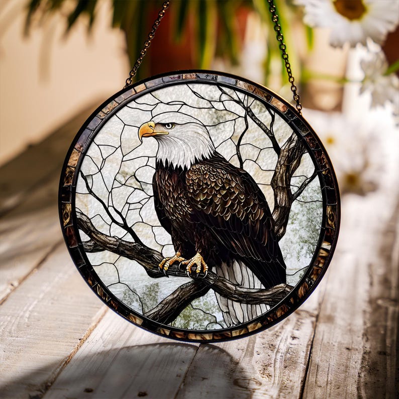 May include: Circular stained glass window depicting a bald eagle on a branch. The eagle has a white head and brown body, with a yellow beak and talons. The background is light with a tree branch design. The window is framed in a dark metal, and is suspended by a chain.