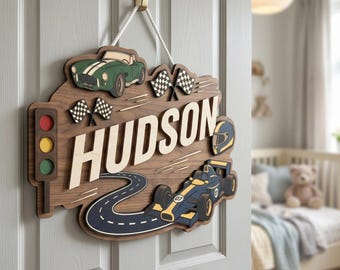 May include: Wooden racing-themed name sign with the name "HUDSON" in large, light-colored letters. The sign features a green classic car, a blue and yellow race car, checkered flags, a helmet, and a traffic light. The sign hangs from a white rope.