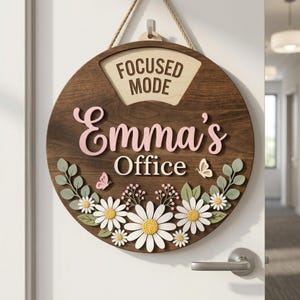 Puede incluir: Un letrero de oficina redondo de madera con las palabras "Focused Mode" y "Emma's Office". El letrero está decorado con margaritas, mariposas y hojas. Cuelga de una cuerda.