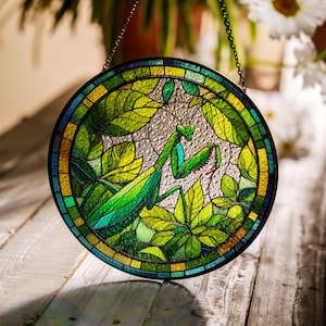 Puede incluir: Una ventana de vidrio coloreado colgante presenta una mantis religiosa verde con una flor roja sobre un fondo de hojas. La ventana es redonda y tiene un marco de metal.
